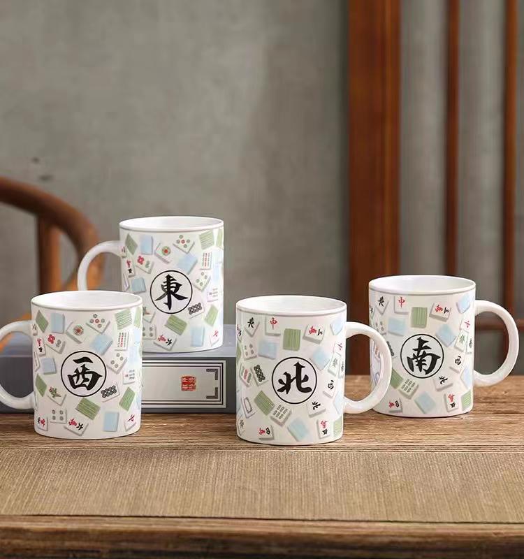 Set of four white mahjong-themed ceramic coffee mugs featuring colorful mahjong tile patterns and black Chinese characters representing East, West, North, and South winds, displayed on a wooden table.