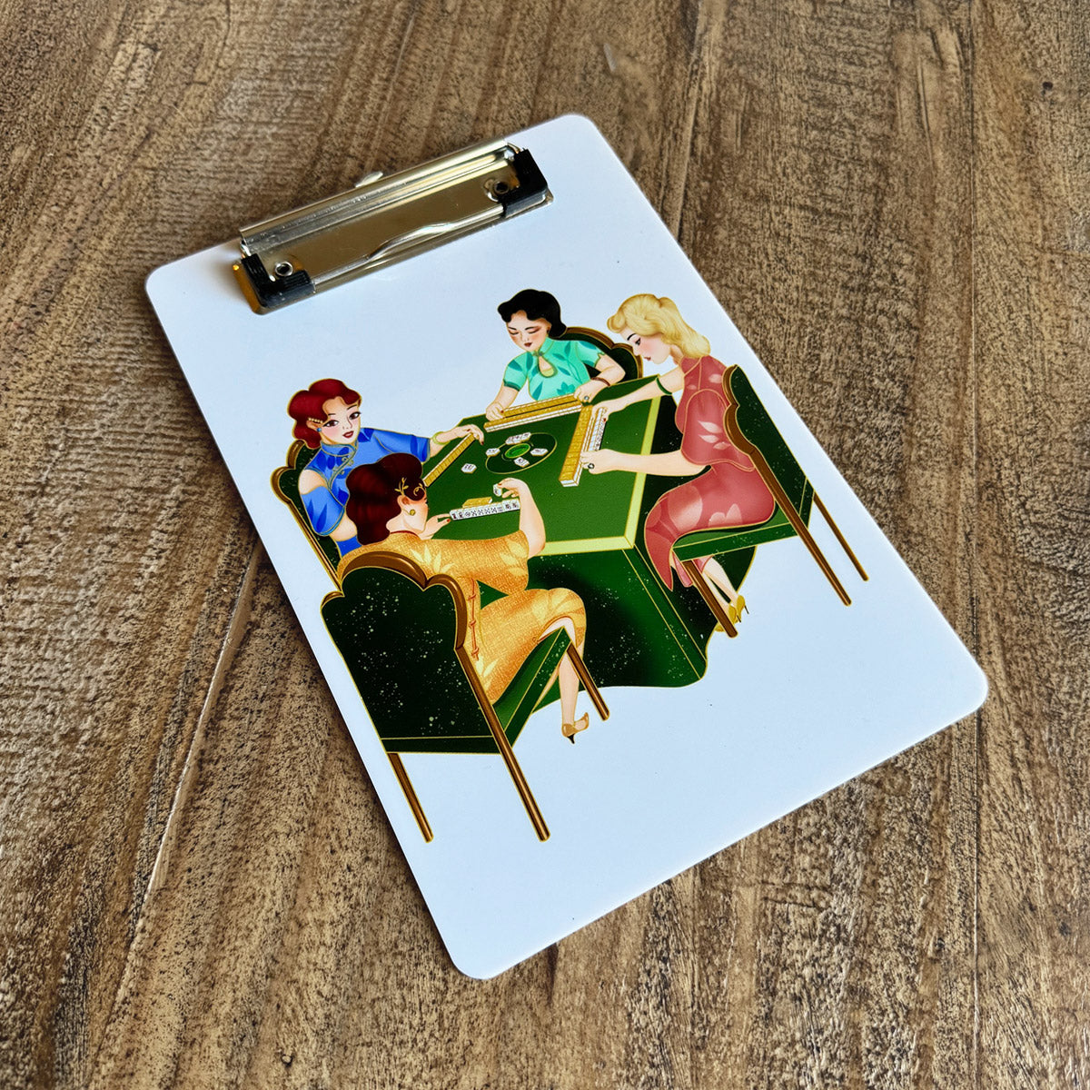 White mahjong clipboard featuring a colorful vintage-style illustration of four women playing mahjong around a green table. Includes a silver metal clip at the top and rests on a rustic wooden surface.