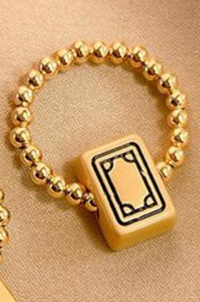Gold beaded mahjong bracelet featuring a cream-colored mahjong tile charm with a black outlined blank tile design, displayed on beige fabric.