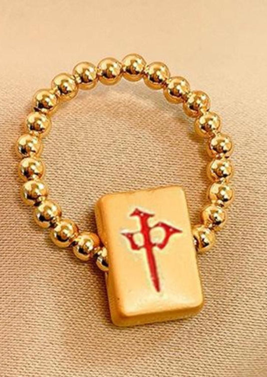 Gold beaded mahjong bracelet featuring a cream-colored mahjong tile charm with a green Chinese character representing the Green Dragon tile, displayed on beige fabric.