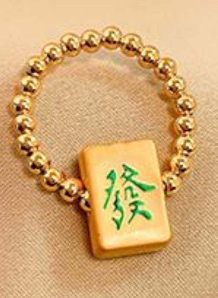 Gold beaded mahjong bracelet featuring a cream-colored mahjong tile charm with a green Chinese character representing the Green Dragon tile, displayed on beige fabric.
