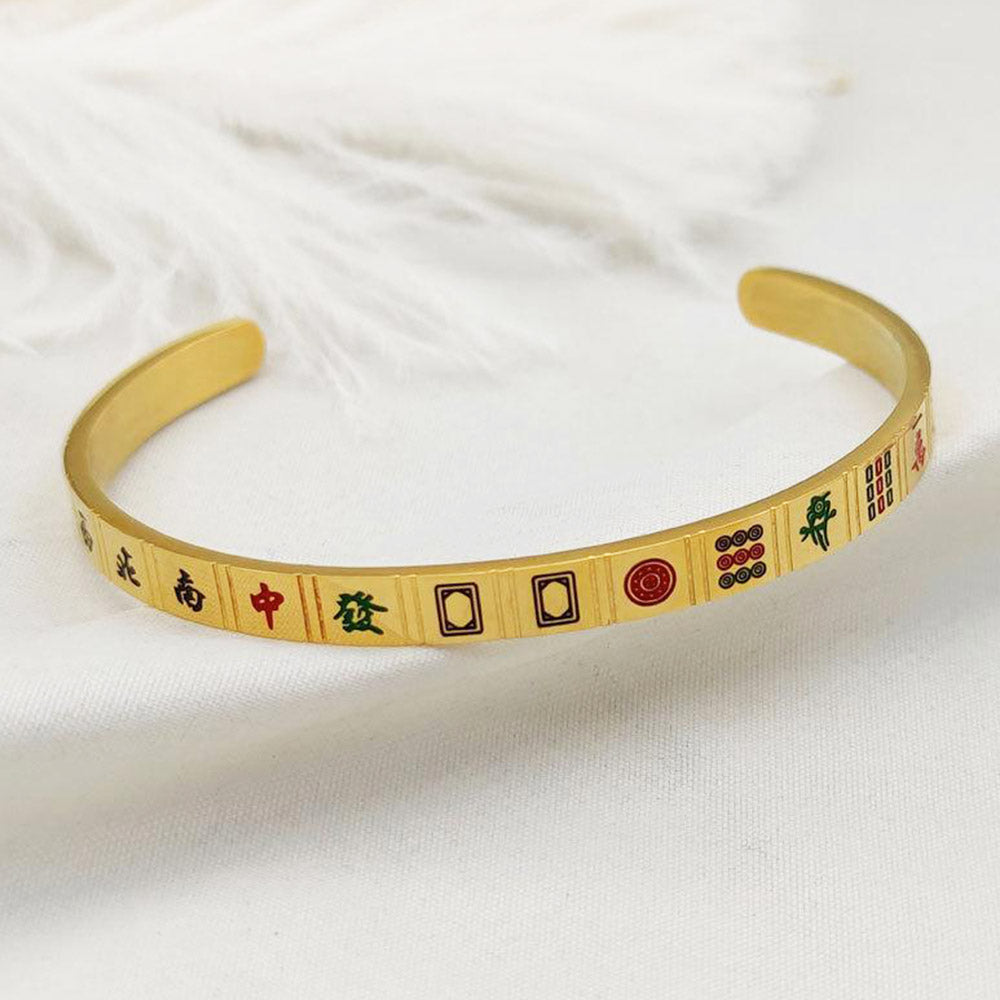 Gold-tone open cuff bracelet featuring colorful mahjong tile symbols, including Chinese characters, bamboo, and circle designs. Elegant mahjong-inspired jewelry displayed on soft white fabric.