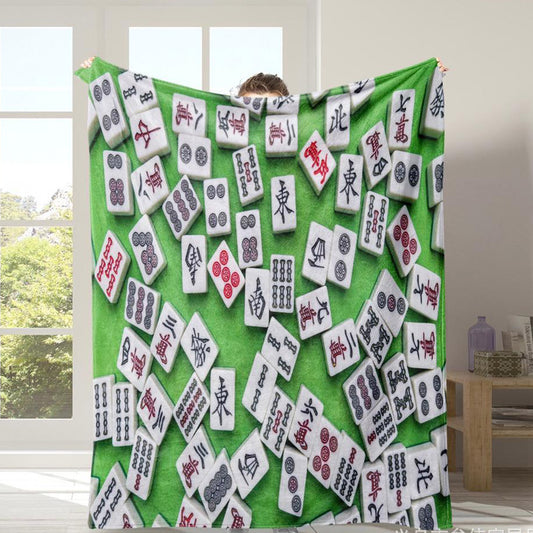 Green mahjong-themed throw blanket featuring a colorful pattern of scattered white mahjong tiles with Chinese characters, bamboo, and circle symbols. Displayed in a bright living room.