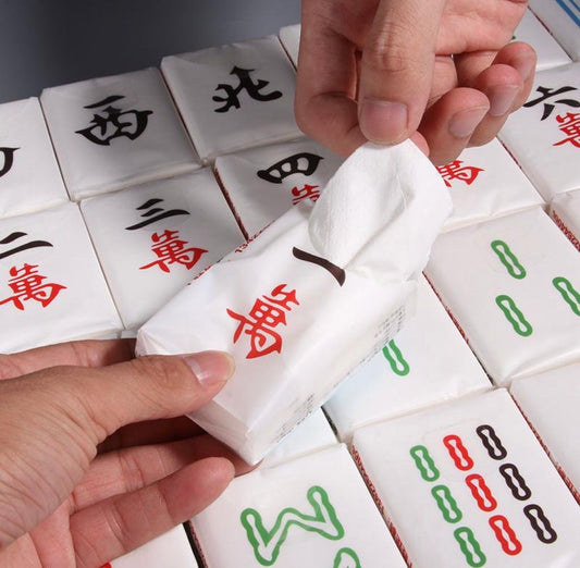Mahjong-themed tissue packs designed to resemble traditional mahjong tiles, featuring Chinese characters and colorful bamboo symbols in red, green, and black, with a hand pulling a white tissue from one pack.