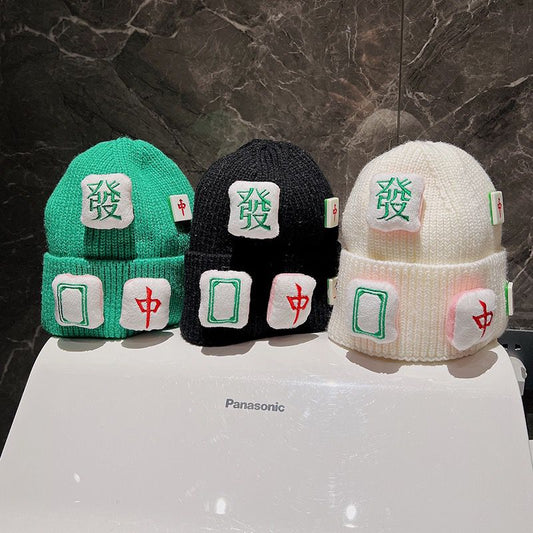 Three mahjong-themed knit beanie hats in green, black, and white, featuring decorative mahjong tile patches with Green Dragon, Red Dragon, and blank tile designs.
