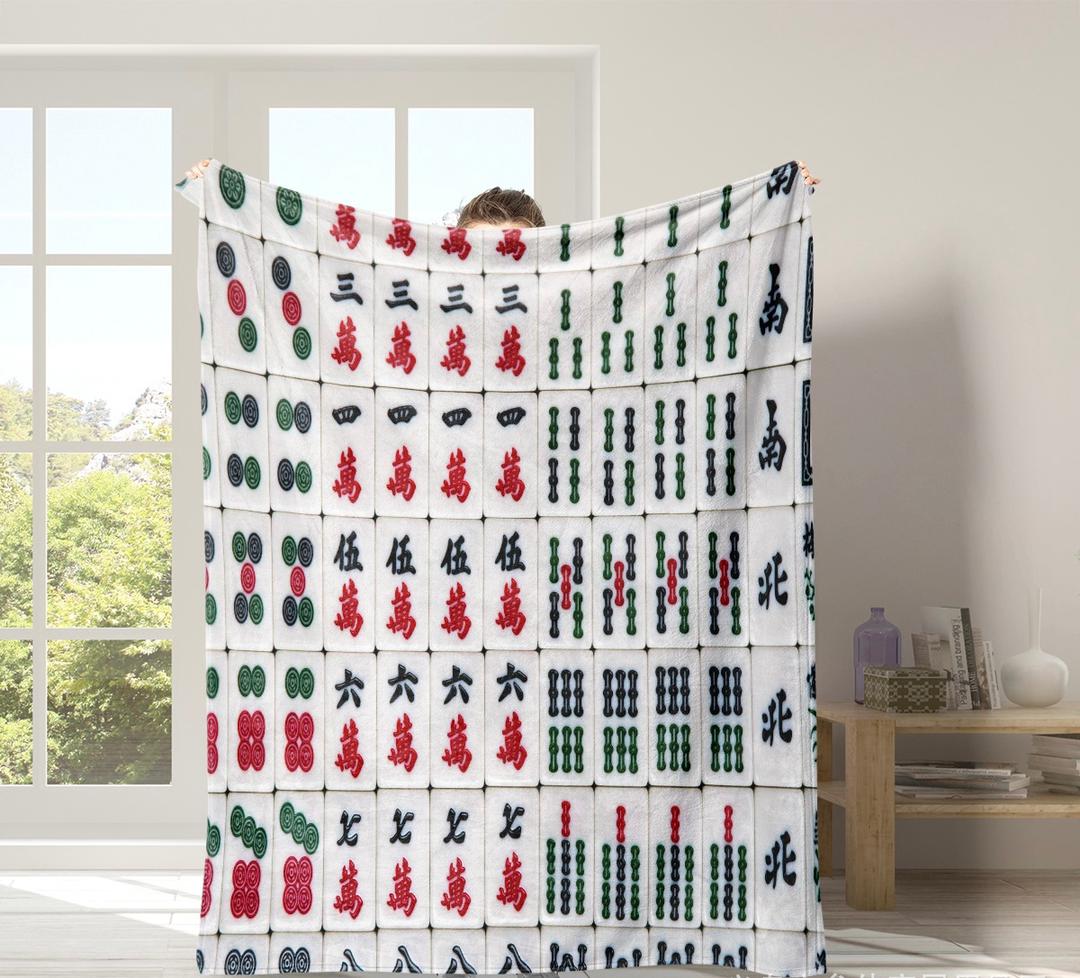 Large white mahjong-themed throw blanket featuring a colorful pattern of traditional mahjong tiles with Chinese characters, bamboo, and dot symbols in red, green, and black, displayed in a bright living room.