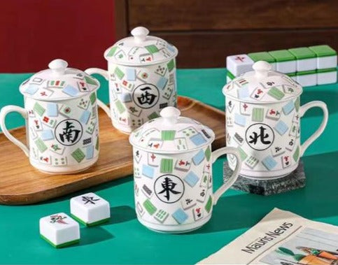 Set of four white mahjong-themed ceramic mugs with matching lids, featuring colorful mahjong tile patterns and Chinese characters representing East, West, North, and South winds, displayed on a green table with mahjong tiles.