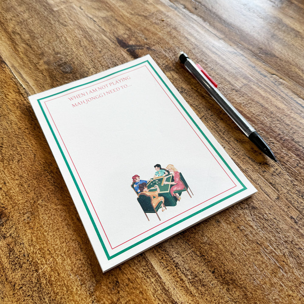Mahjong-themed to-do list notepad featuring the phrase "When I Am Not Playing Mah Jongg I Need To..." with a green and red border and a vintage illustration of four women playing mahjong. Displayed on a wooden table beside a pencil.