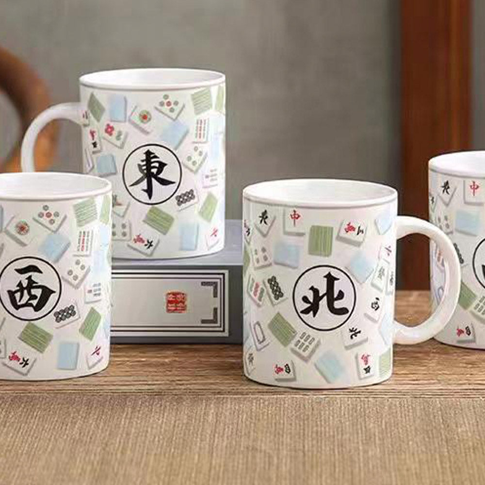 Close-up of white mahjong-themed ceramic coffee mugs featuring colorful mahjong tile patterns and black Chinese characters representing East, West, and North winds, displayed on a wooden table.