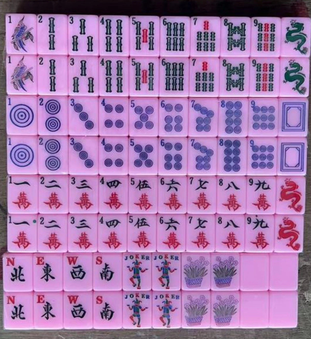 Complete pink American mahjong tile set arranged in rows, featuring colorful bamboo, dots, Chinese characters, dragons, winds, flower tiles, and jokers with traditional designs.