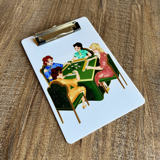 White mahjong clipboard featuring a colorful vintage-style illustration of four women playing mahjong around a green table. Includes a silver metal clip at the top and rests on a rustic wooden surface.