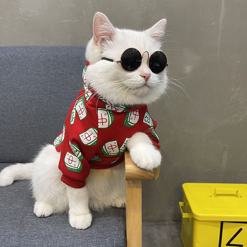 White cat wearing round black sunglasses and a red mahjong-themed sweater featuring colorful mahjong tile patterns in red, green, and white, sitting on a gray chair.